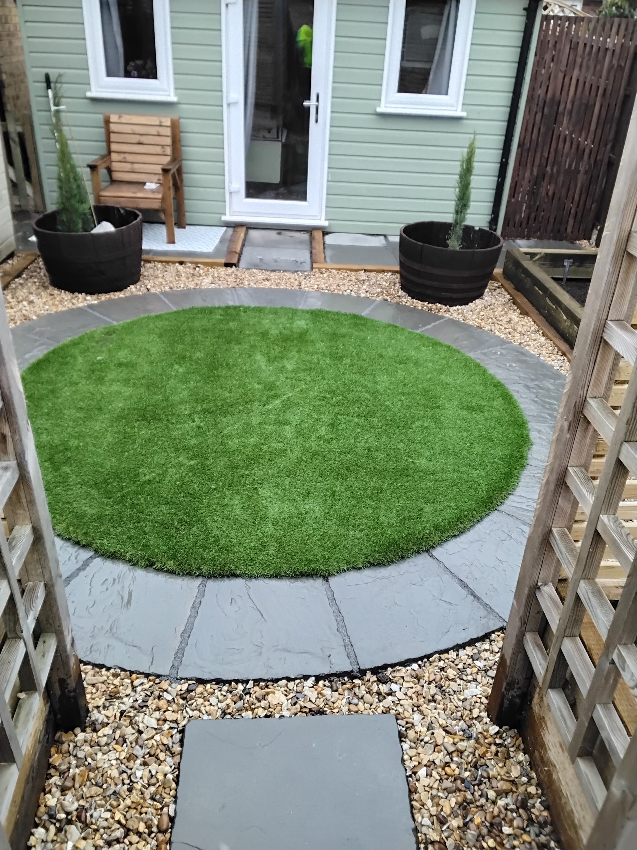 A circular artificial lawn edged with slate, set in gravel in front of a garden room